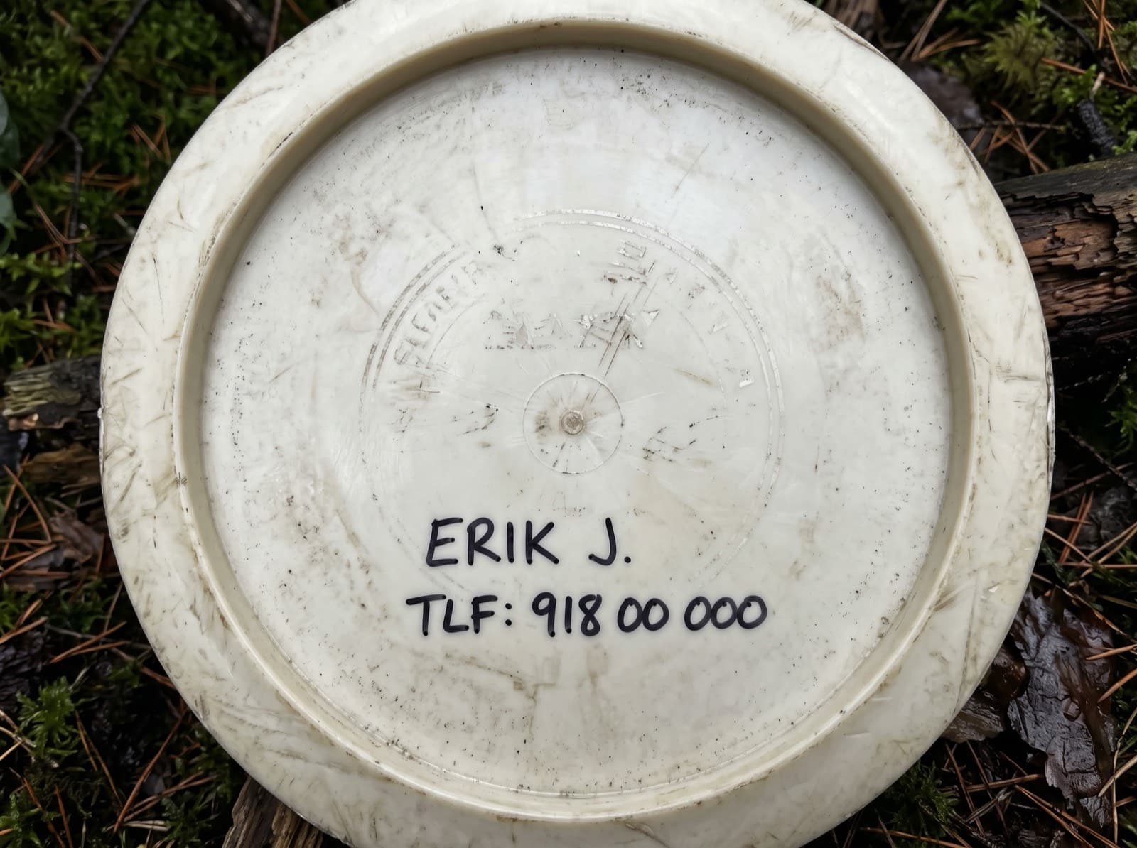 Undersiden av en slitt, hvit disk som ligger i mose, med fornavn og telefonnummer skrevet på med svart tusj